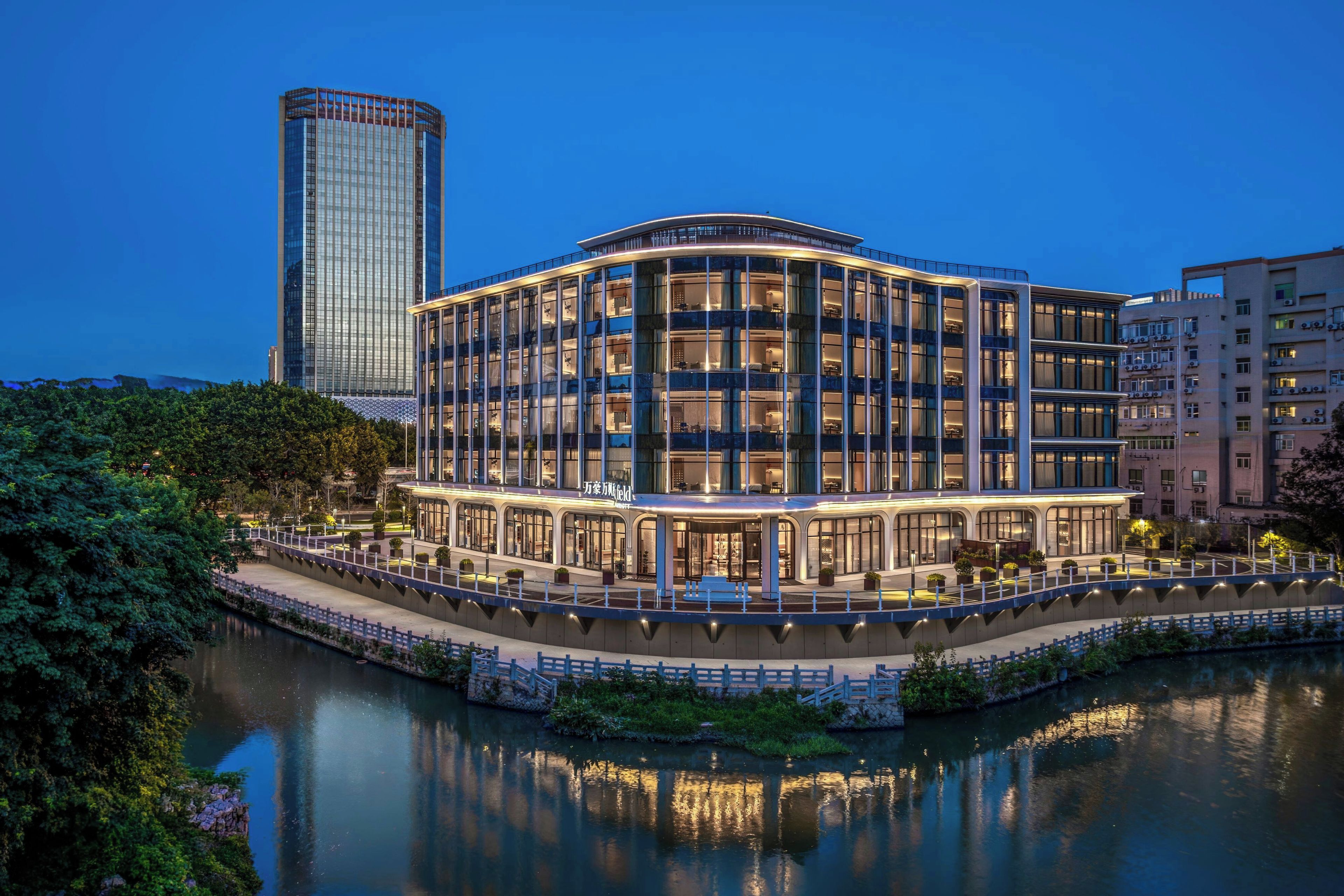 A multi-story hotel with a glass facade, located beside a body of water, with a high-rise building in the background.