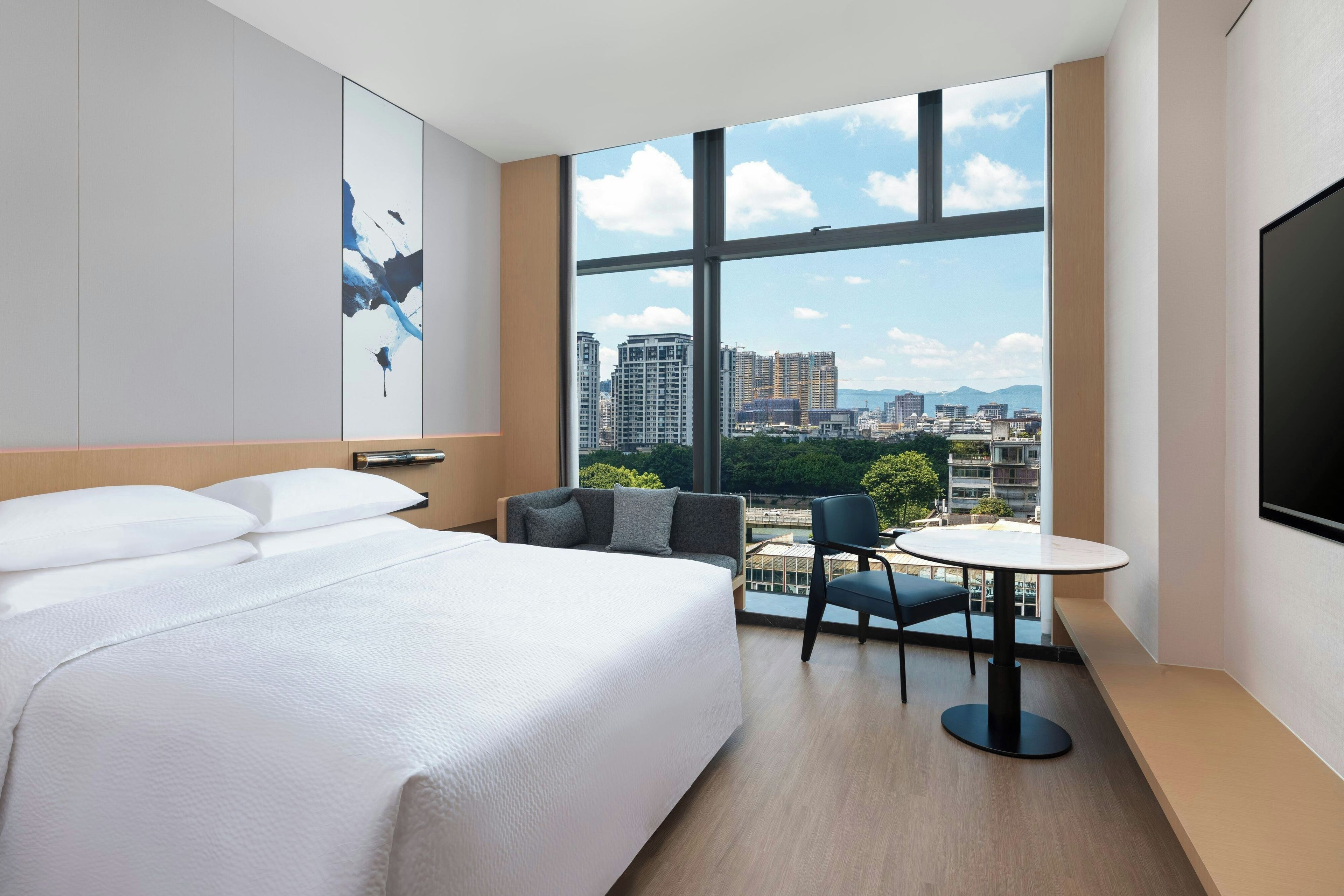 Habitación de hotel con una cama grande, una mesita pequeña y una silla, un ventanal con vistas a la ciudad y un televisor de pantalla plana montado en la pared.