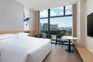 Une chambre d’hôtel avec un grand lit, une petite table et une chaise, une grande fenêtre donnant sur la ville et un téléviseur à écran plat fixé au mur.