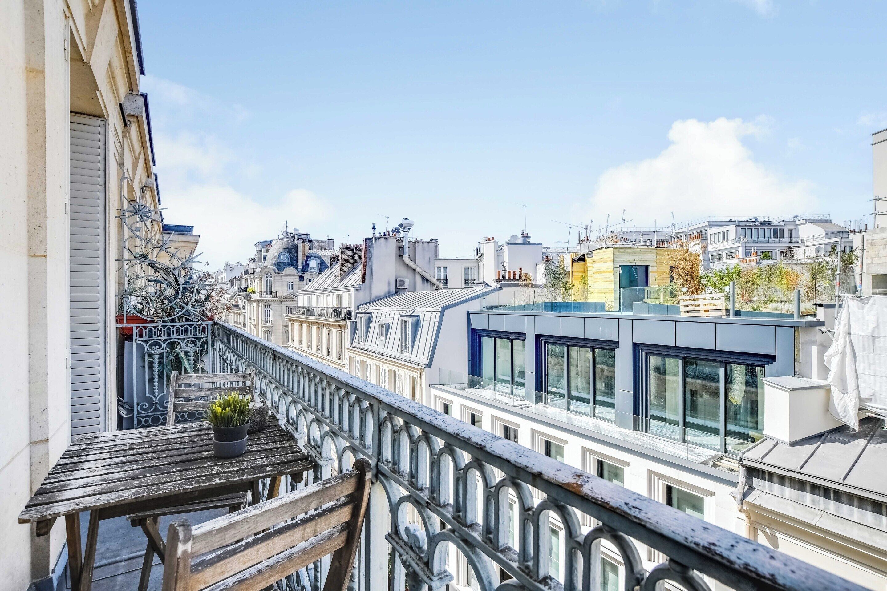 A balcony with a wooden table and potted plant, overlooking a cityscape with various buildings.