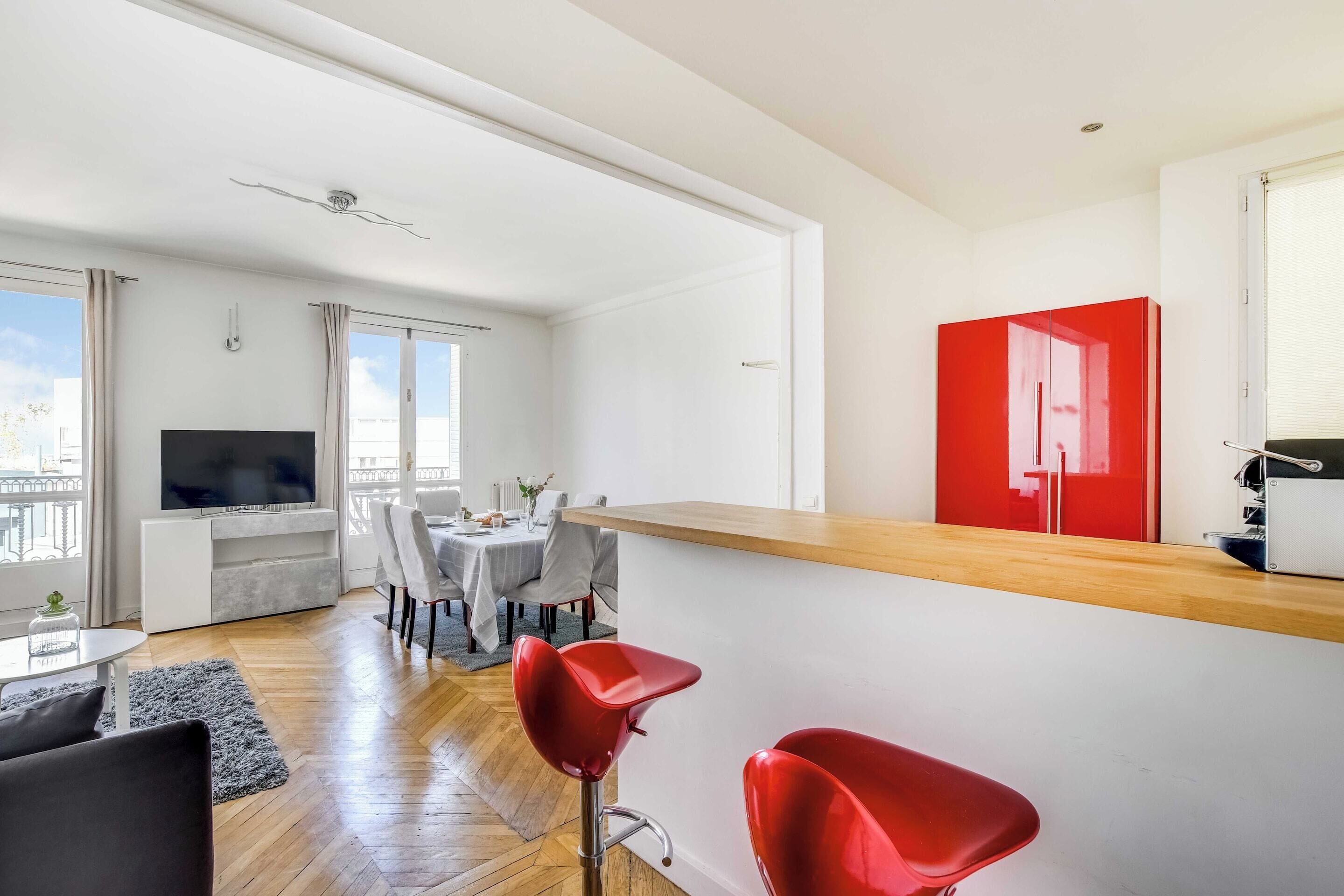 A modern living space with a dining area, a kitchen with a red cabinet, and a flat-screen TV.