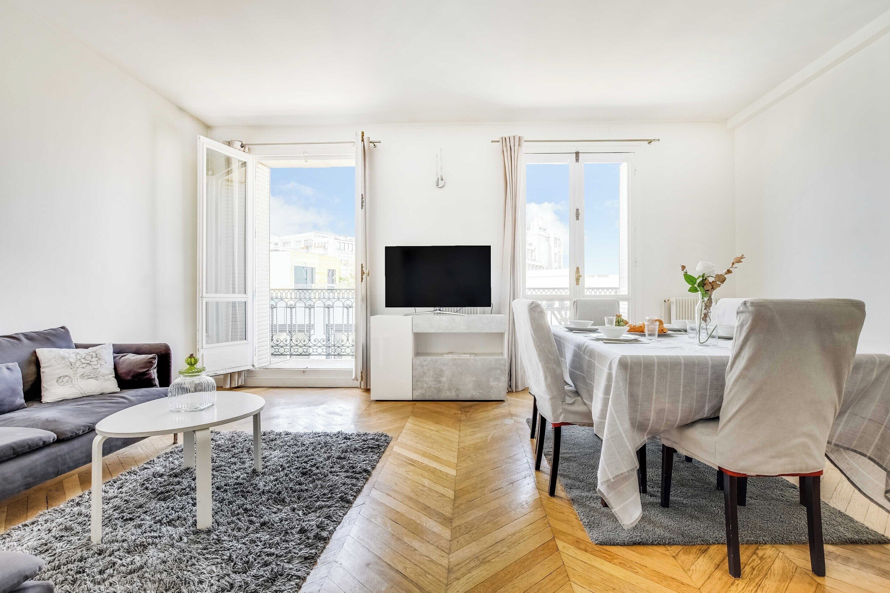 A modern living room with a sofa, a dining table with chairs, a TV, and a view of the city through open doors.