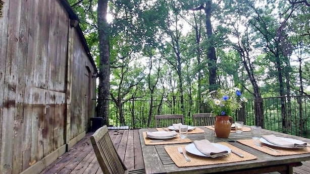 Une terrasse en bois aménagée à l’extérieur, avec une table dressée pour le repas, entourée d’arbres.