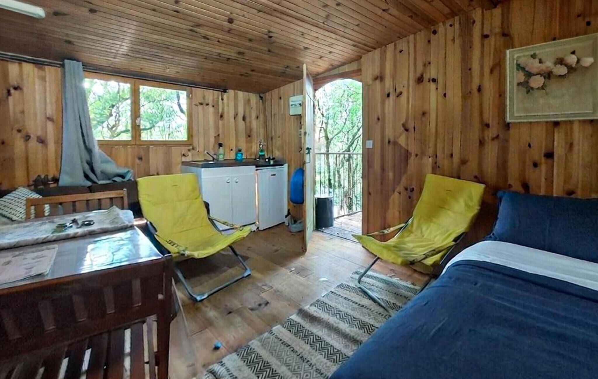 Una cabaña de madera con una cama, dos sillas amarillas, una pequeña cocina y vistas a árboles.