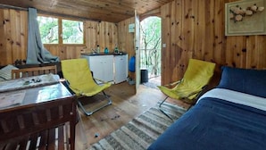 Une cabane en bois avec un lit, deux chaises jaunes, une petite kitchenette et une vue sur les arbres.