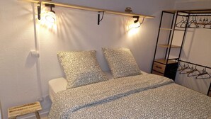 A bedroom with a bed, two pillows, a bedside table, a small stool, a hanging light fixture, and a clothing rack. - The Dune Jewel Box • Vendres-Plage • Sea & Pool (Vendres)