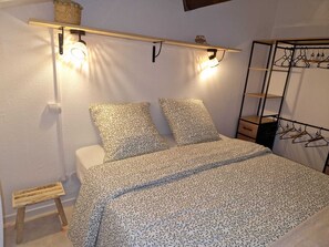 Ein Schlafzimmer mit einem Bett, zwei Kissen, einem Nachttisch, einem kleinen Hocker, einer Pendelleuchte und einem Kleiderständer.