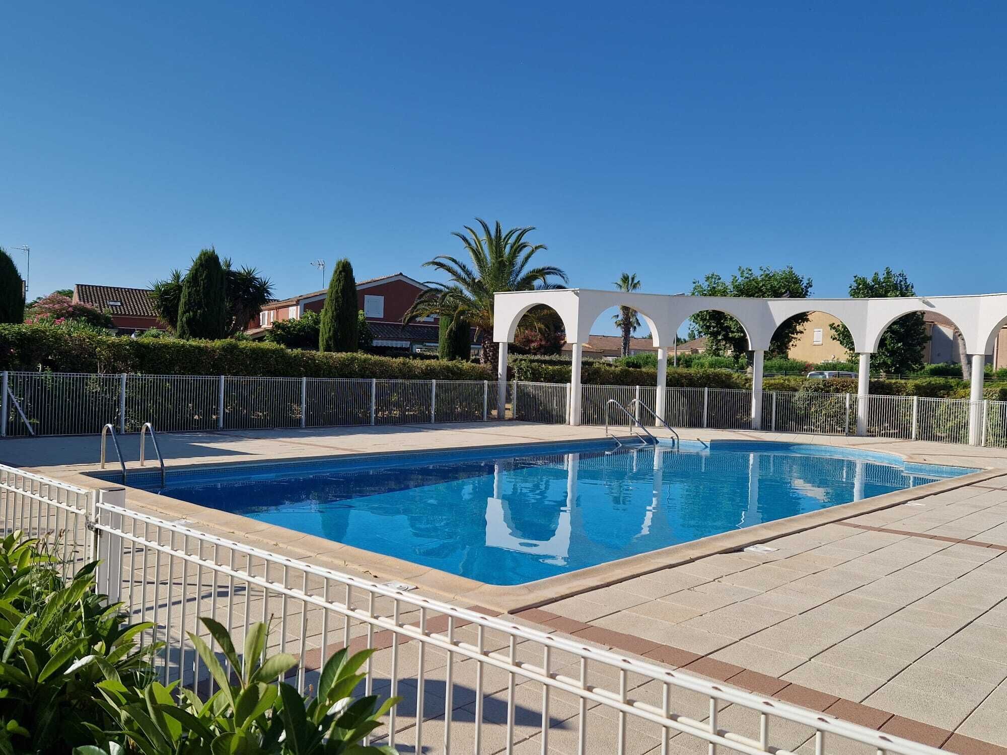 Una piscina con valla blanca, una estructura de pérgola y edificios residenciales al fondo.