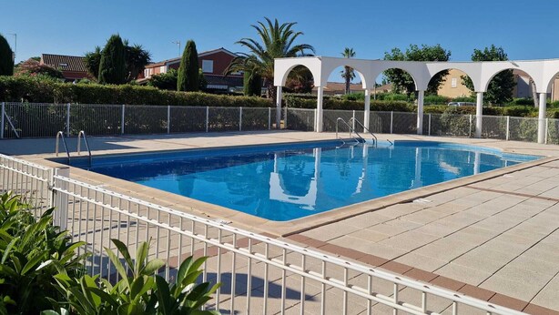 A swimming pool with a white fence, a pergola structure, and residential buildings in the background. - The Dune Jewel Box • Vendres-Plage • Sea & Pool (Vendres)