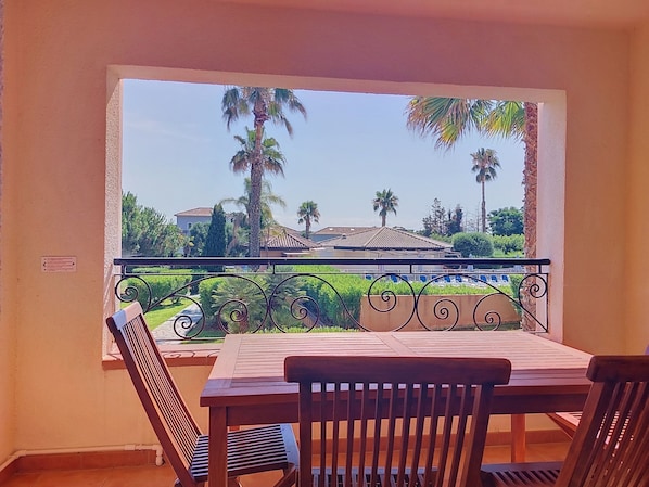 A balcony with a table and chairs, overlooking a garden with palm trees and a building. - Alba & Tramonto by Interhome (Bravone)