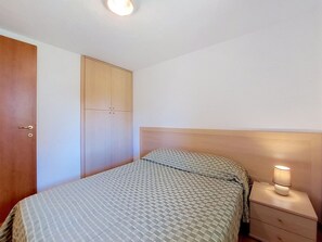 Un dormitori amb llit, una tauleta de nit amb una làmpada, un armari i una porta.