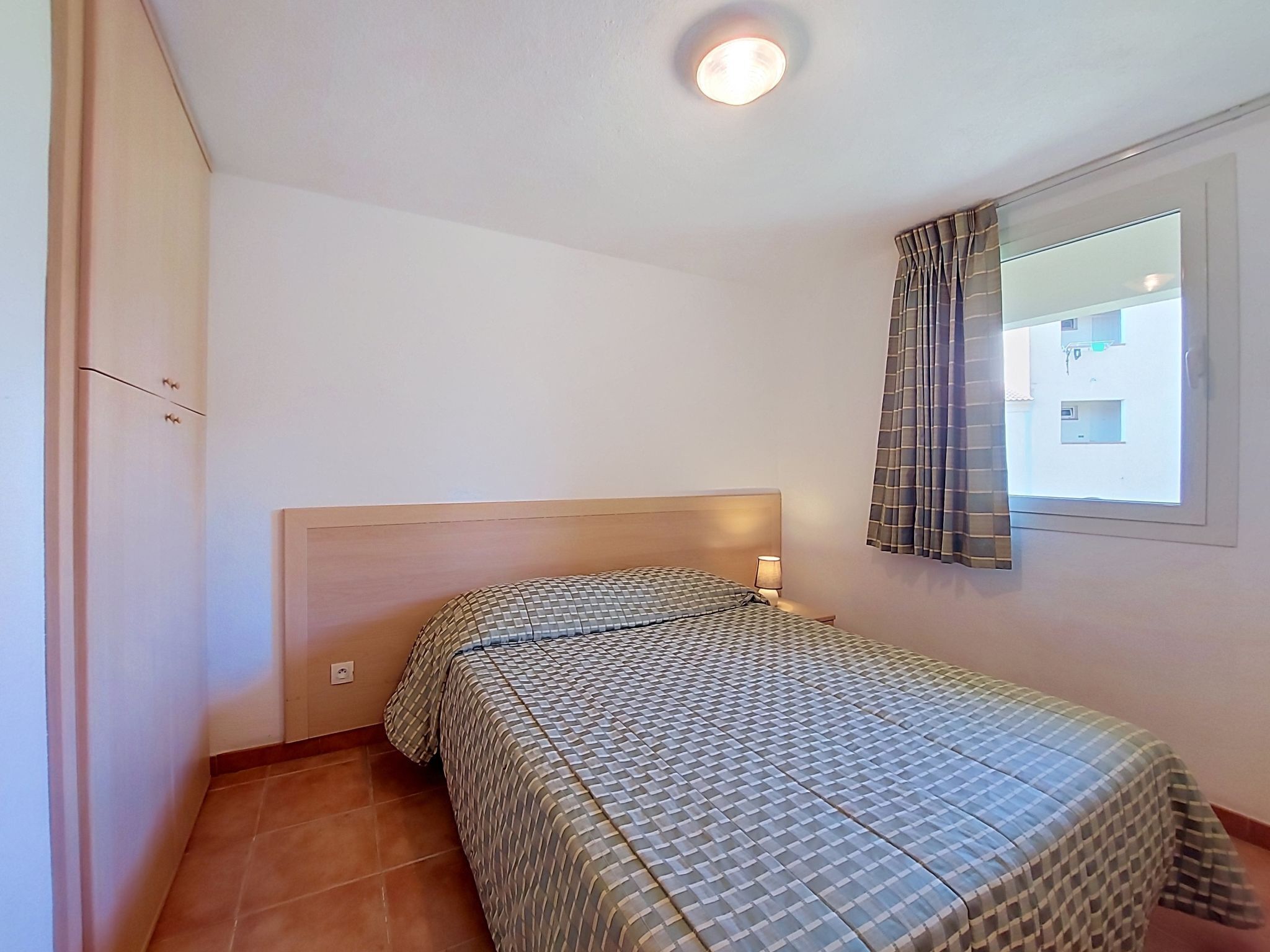 Ein Schlafzimmer mit einem Einzelbett, einem Nachttisch, einer Wandlampe, einem Fenster mit Vorhängen und einer Tür mit Griff.