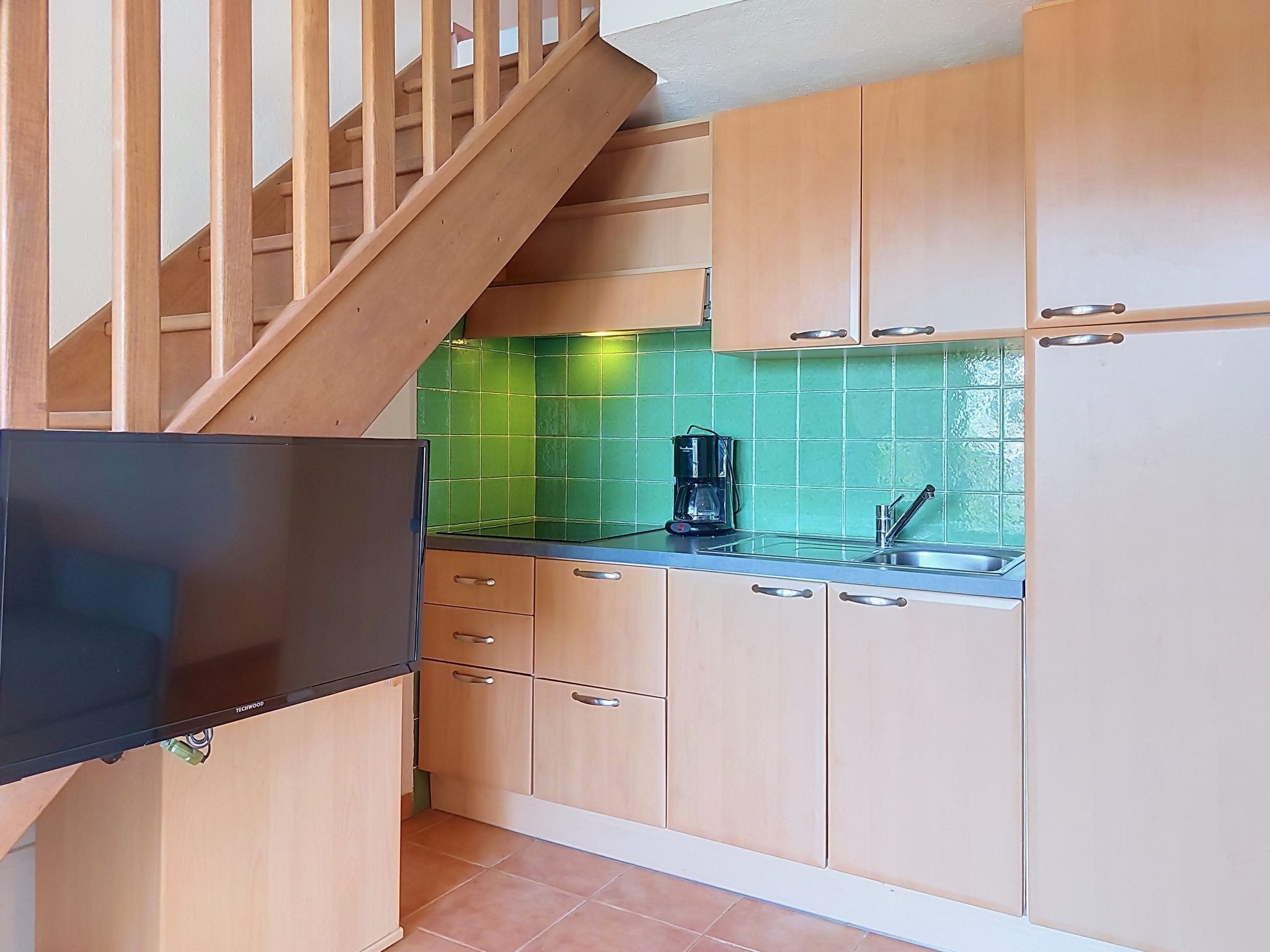 Et køkken med træskabe, et sort køleskab, et grønt flisebaggrund og en trætrap.