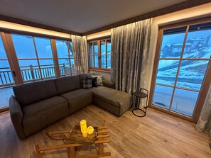 A cozy living room with a sectional sofa, a small table with yellow cups, and large windows offering a view of a snowy landscape. - Chalet d'Mot, (Samnaun-Ravaisch) by Interhome (Samnaun)