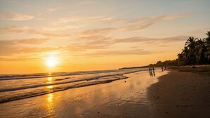 A beach with the sun setting over the horizon, waves gently lapping the shore, and a few people walking along the beach. - Uvita vacation rental with private pool, rancho lounge, and lush garden just minutes from the beach. (Uvita)