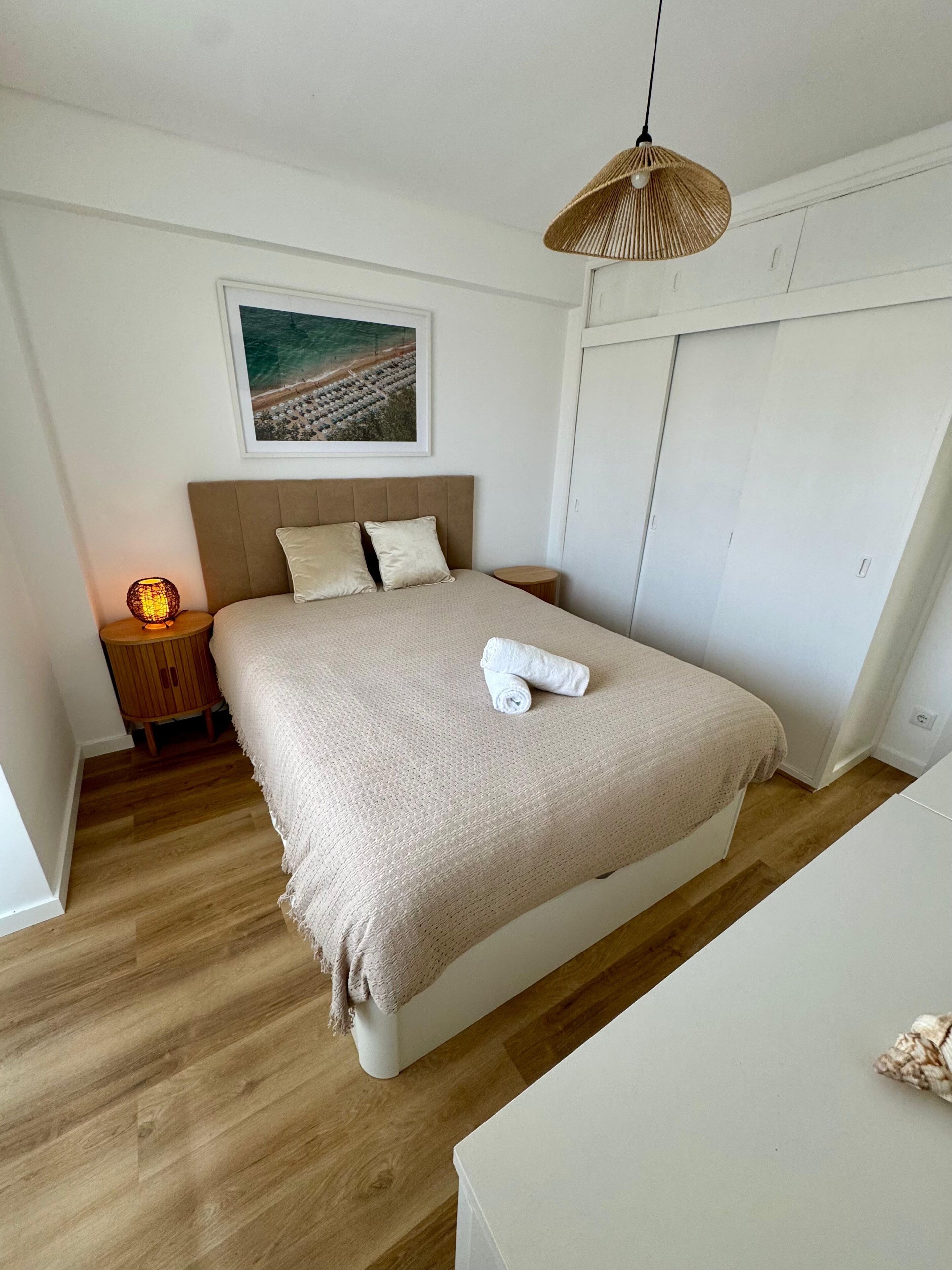 A bedroom with a bed, a nightstand, a lamp, a hanging light, a framed picture on the wall, and a wardrobe.