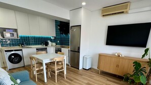 Ein modernes Wohnzimmer mit Sofa, Esstisch mit Stühlen, einer Küchenzeile inklusive Kühlschrank und einem an der Wand montierten Fernseher.