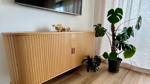 Ein Holzschrank, darüber ein Flachbildfernseher, ein Topfpflanze und ein Fenster mit Vorhängen.