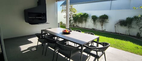 Un patio al aire libre con mesa y sillas, una parrilla negra y vistas a un jardín cuidado.