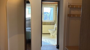 Ein Flur mit Holzboden, weißen Wänden und einem Oberlicht. Zwei Blumentöpfe und ein weißer Metallregal sind ebenfalls vorhanden.