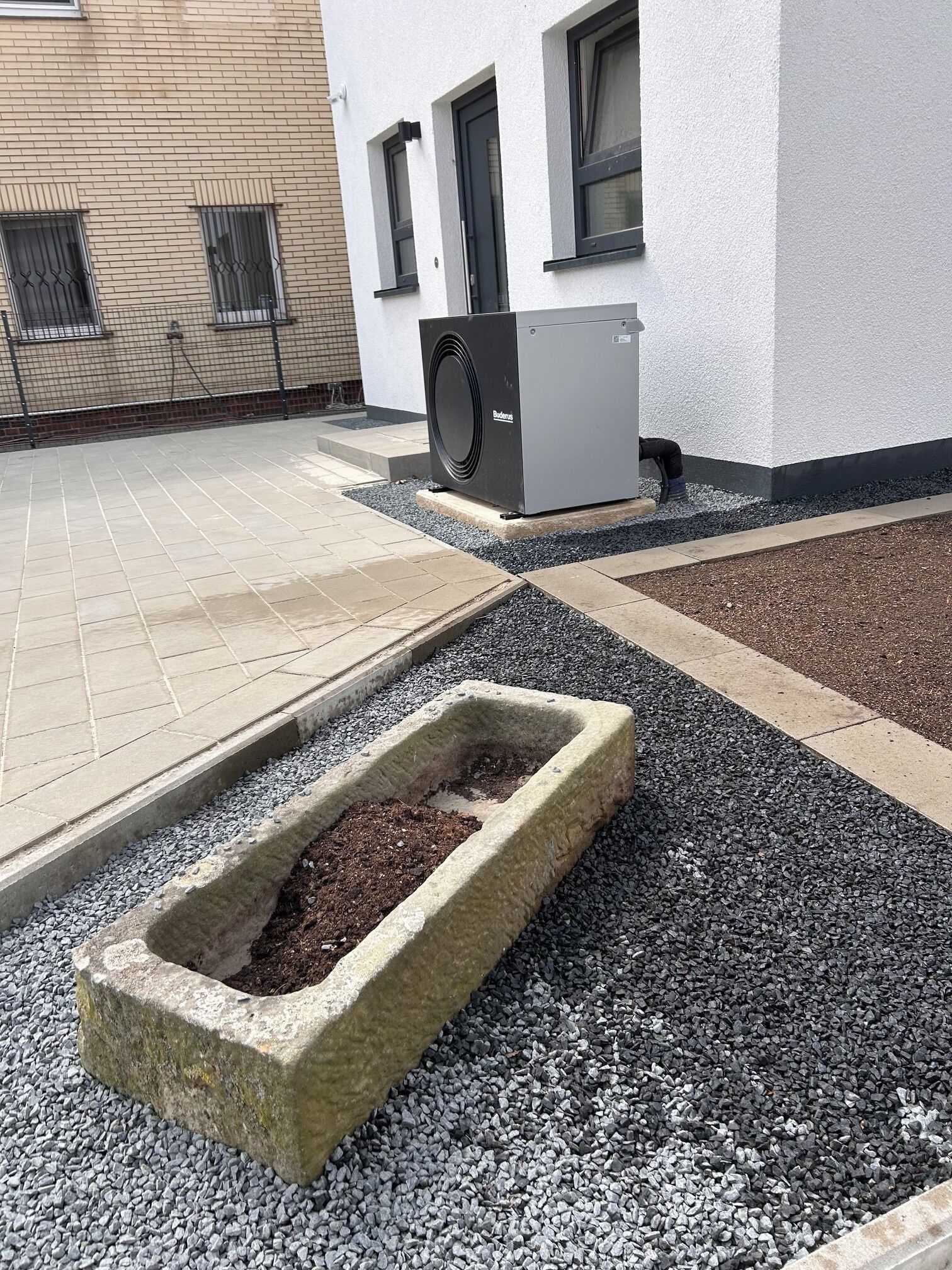A building exterior with a paved area, a rectangular planter with soil, and a large air conditioning unit.