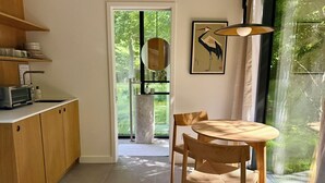 Een keuken met houten kasten, een eettafel met stoelen, een groot raam met uitzicht op groen en een kunstwerk in een lijst aan de muur.