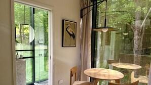 Een kamer met een schuifdeur naar een balkon, een ronde houten tafel met stoelen, een schilderij in een lijst aan de muur en een hanglamp.