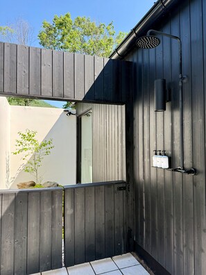 Ein moderner, überdachter Duschenbereich im Freien mit Holzlattenzaun, einer Metallbrause und einem an der Wand befestigten Seifenspender.