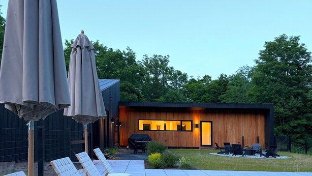 Un'area salotto esterna moderna con poltrone relax e un braciere, un edificio in legno con ampie finestre, e un cielo sereno.