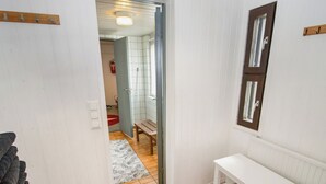 Un baño con paredes blancas, un banco de madera, un tapete rojo y una puerta que da a otra habitación.