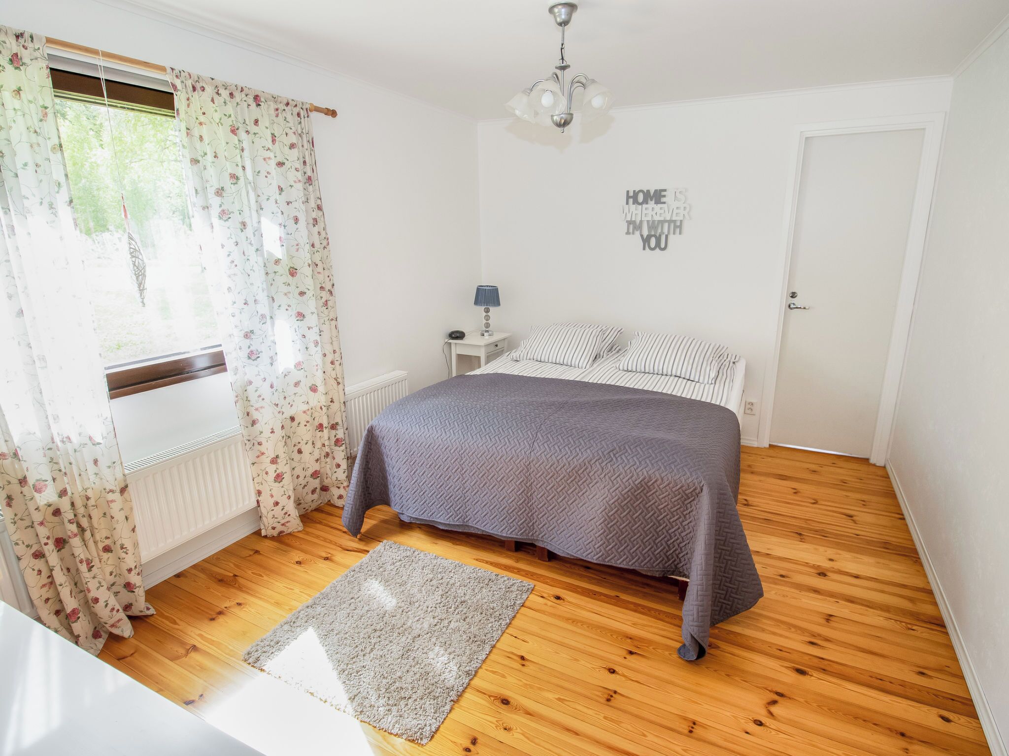 Un dormitorio con cama, mesita de noche, lámpara, radiador, ventana con cortinas florales y una puerta.