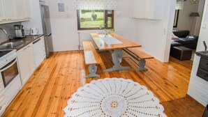 Una cocina con suelo de madera, armarios blancos, una mesa de madera, un banco, una araña y vistas al exterior con vegetación.