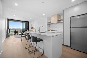 A modern kitchen with a large island, bar stools, a stainless steel refrigerator, and a view of the city through large windows. - CASSA CASINO TOWER 2 - Brand New Premium Residence Managed by The Cassa (Broadbeach)