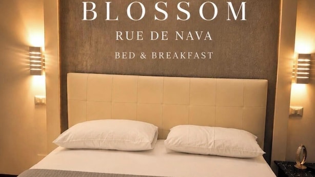 A hotel room with a large bed, two pillows, and a headboard with the hotel's name and location. There are two bedside lamps and a small table with a ring on it. - Blossom Rue De Nava B&B (Reggio Calabria)