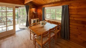 Un interior de madera con una mesa de comedor, sillas, un florero con flores y vistas a través de las ventanas.