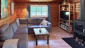 Interior acogedor de una cabaña con un sofá seccional, una mesita con una vela, un televisor y una estantería.