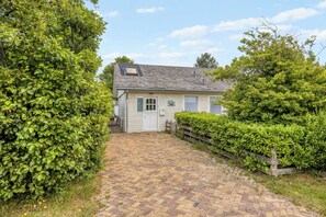 Een huis met een verharde oprit die uitkomt bij een witte deur, omringd door weelderige begroeiing.