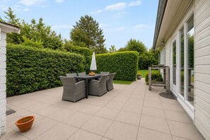 Een terras met een tafel en stoelen, omringd door een haag en een gebouw met grote ramen.