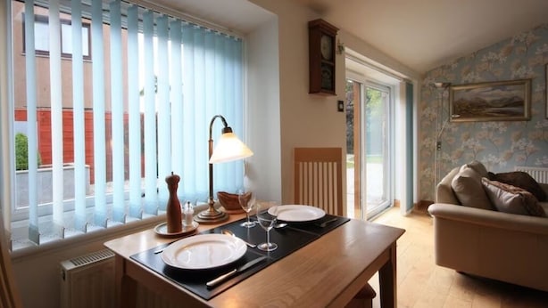 A dining area with a table set for two, a window with blinds, a lamp, and a view of the outside.