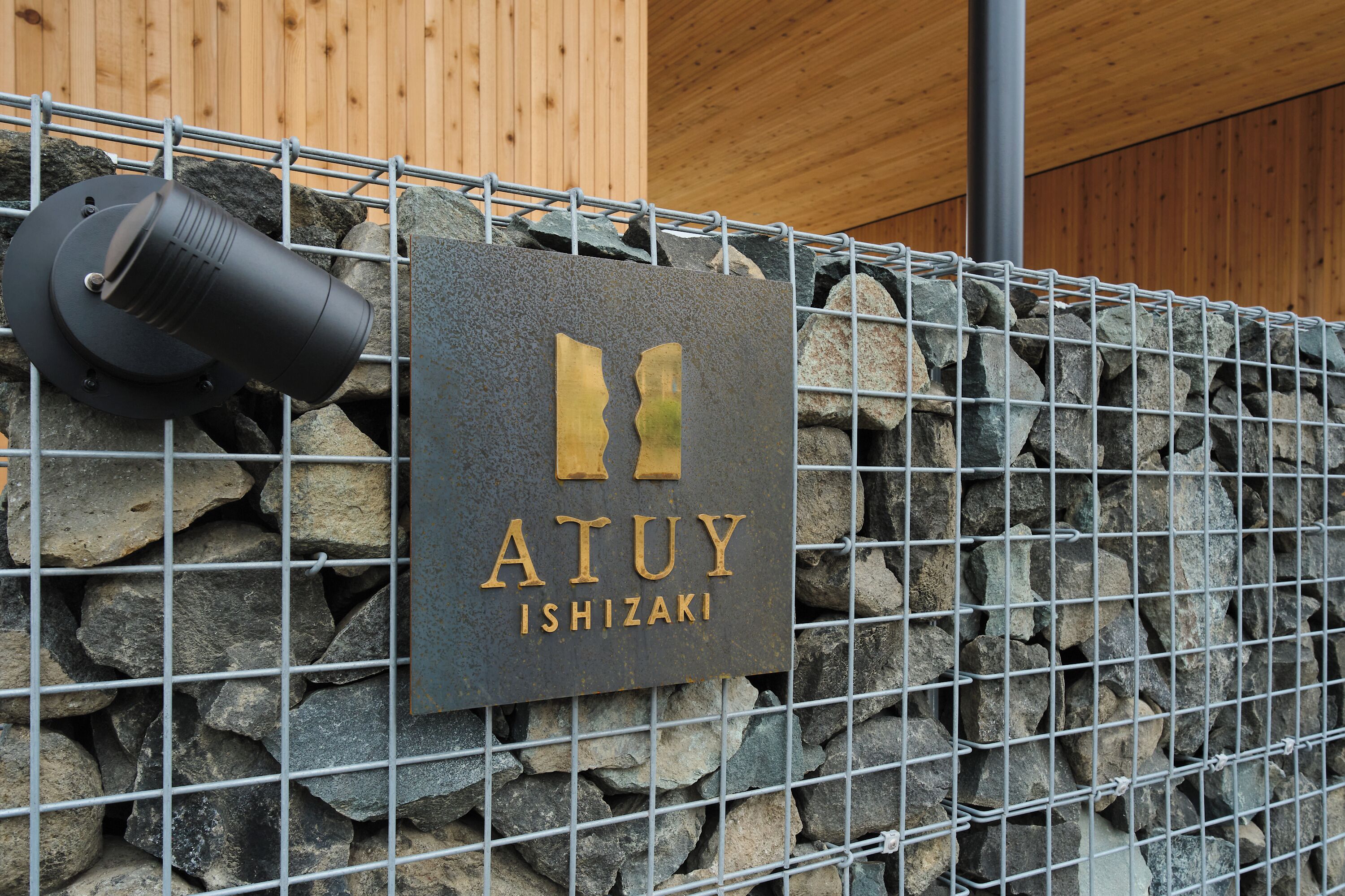 A stone wall with a metal fence, a sign with 'ATUY ISHIZAKI' text, and a cylindrical object on the fence.