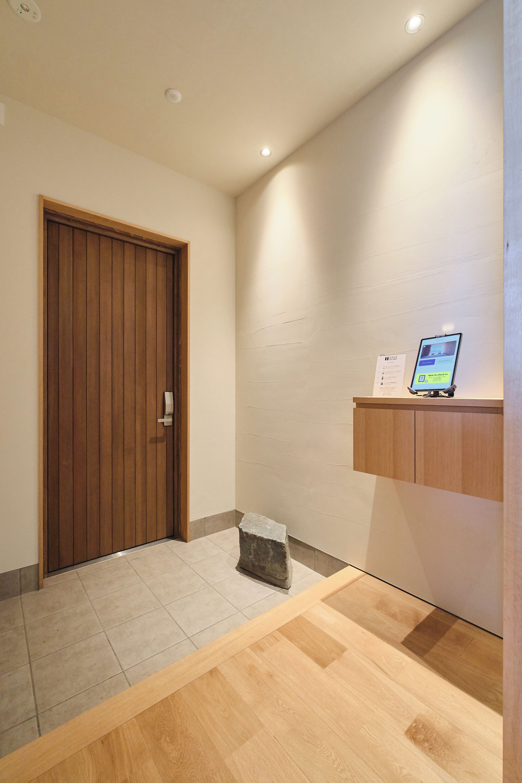A hallway with a wooden door, a stone slab, a wooden floor, and a wall-mounted shelf with a tablet.