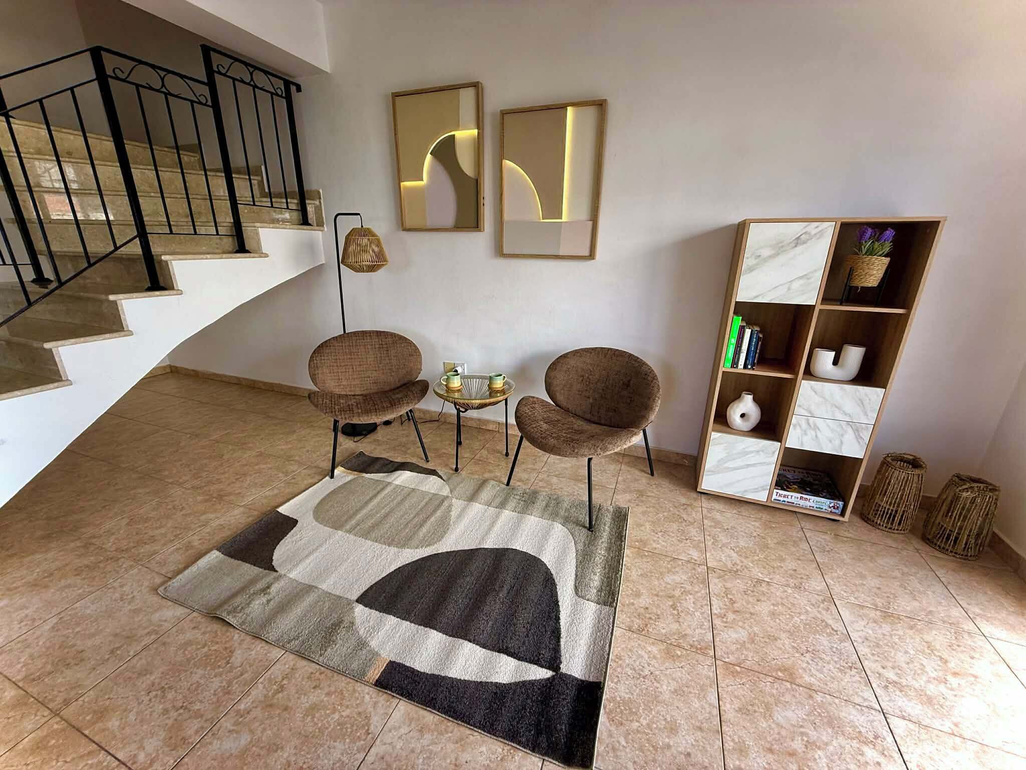 A modern living room with a staircase, two armchairs, a coffee table, a rug, and a bookshelf.