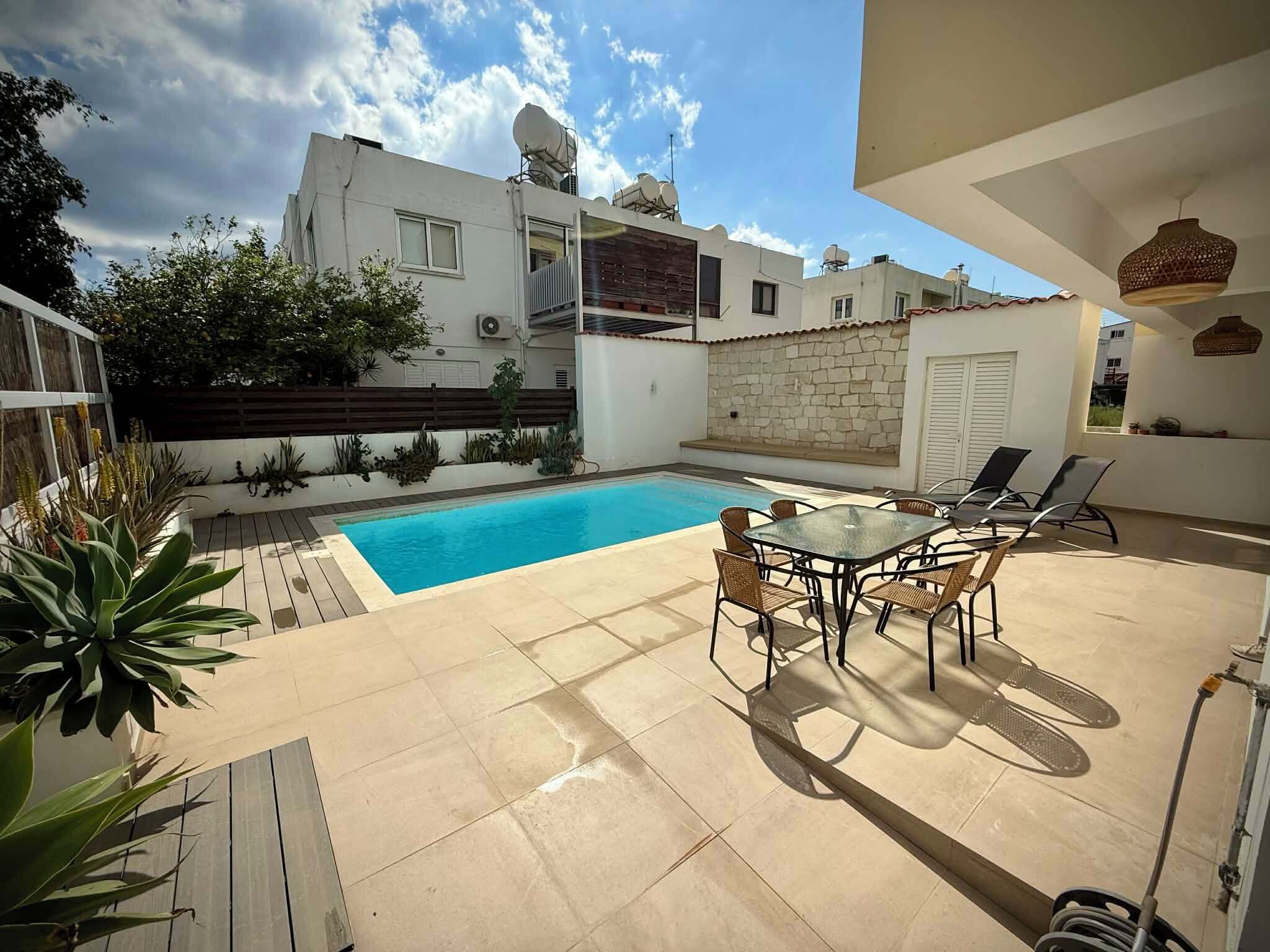 A swimming pool with a patio area, including a table and chairs, surrounded by plants and a stone wall.