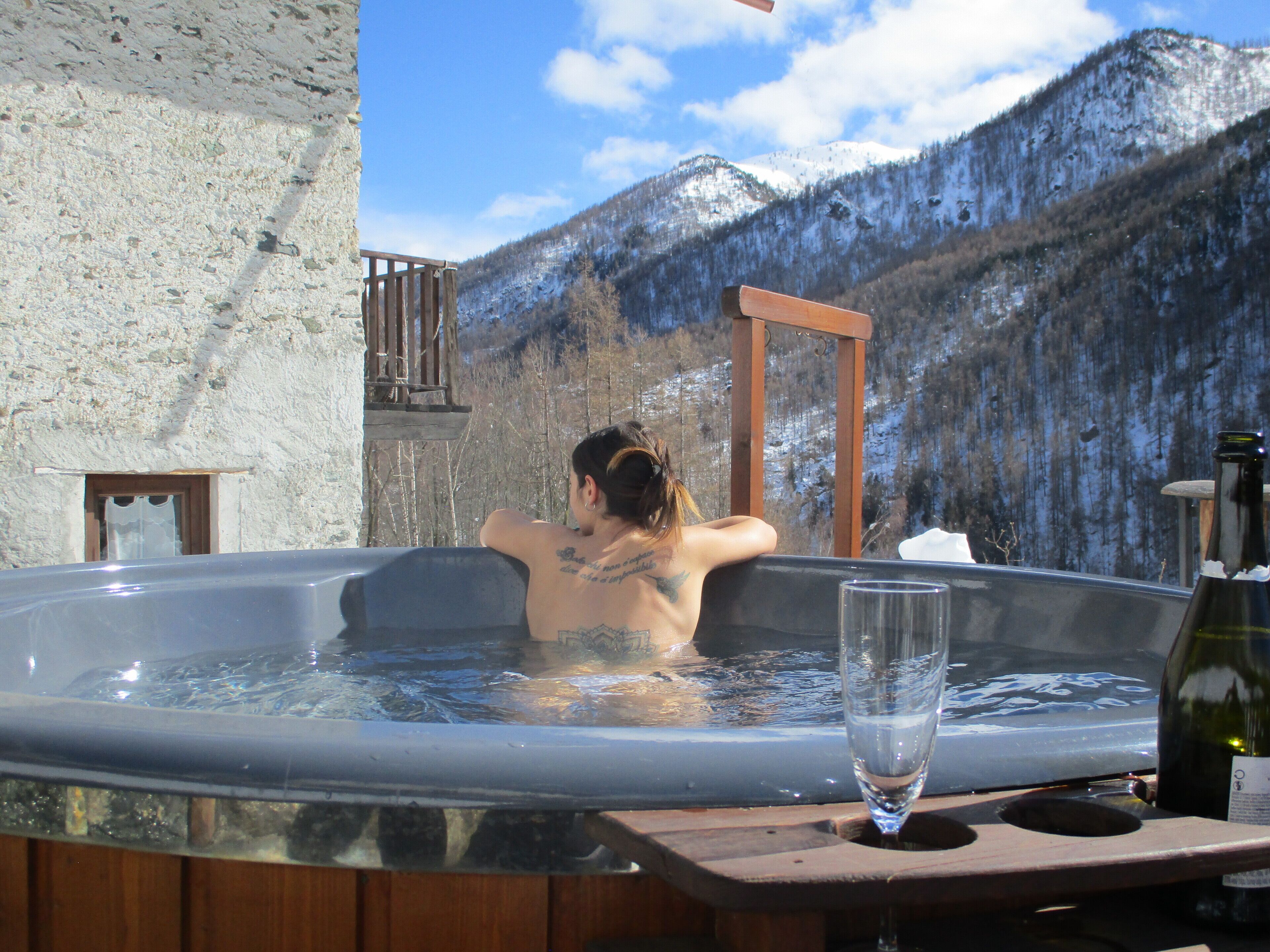 Una persona en un jacuzzi con vista a la montaña, una terraza de madera y una botella de vino al lado.