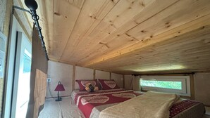 A bedroom with a wooden ceiling, a bed with a red and white bedspread, a window with a view, and a lamp on the floor. - Hempcrete Tiny House (Malbrans)