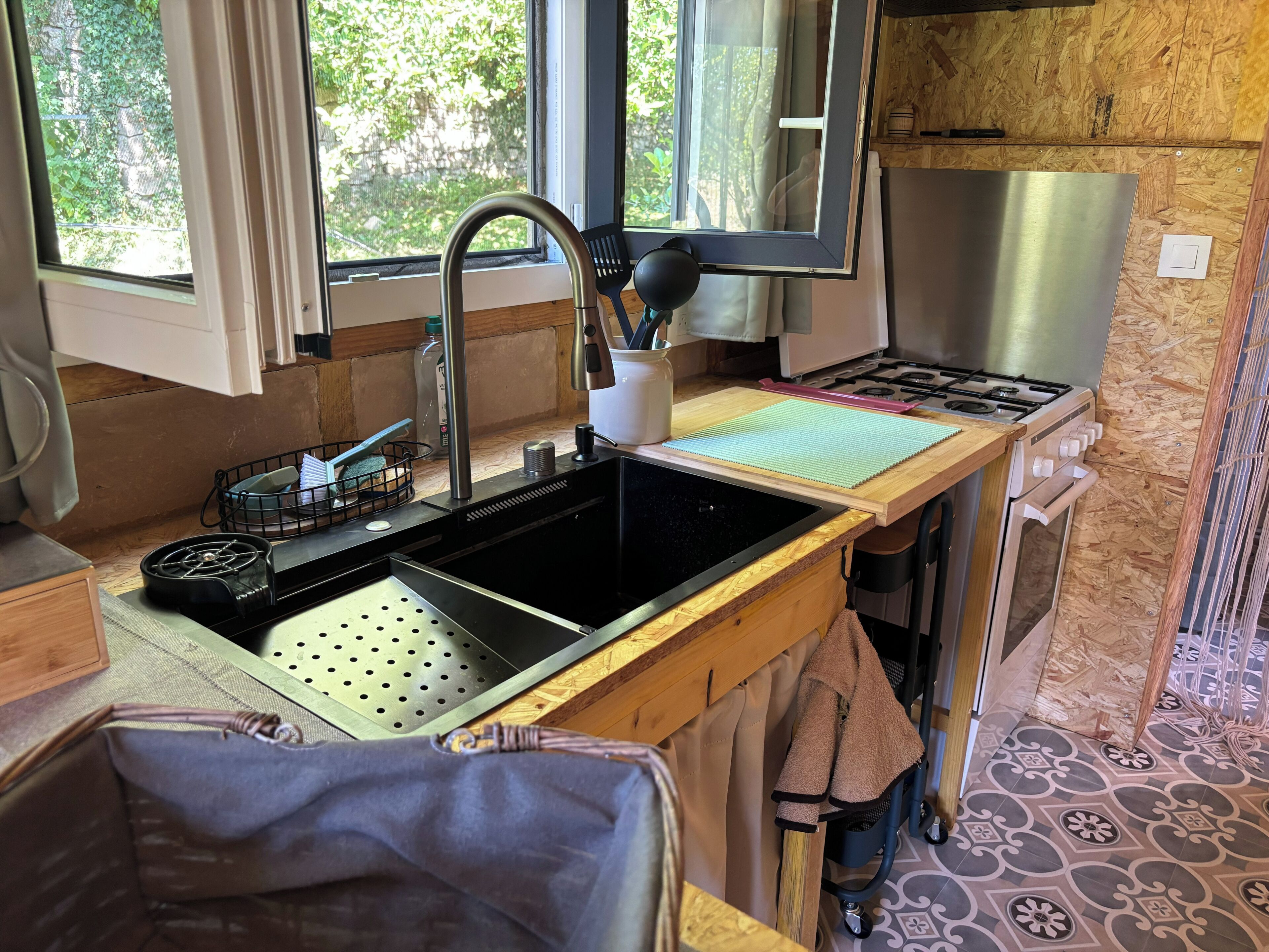 A kitchen with a stainless steel sink, a stove, and a window overlooking greenery.