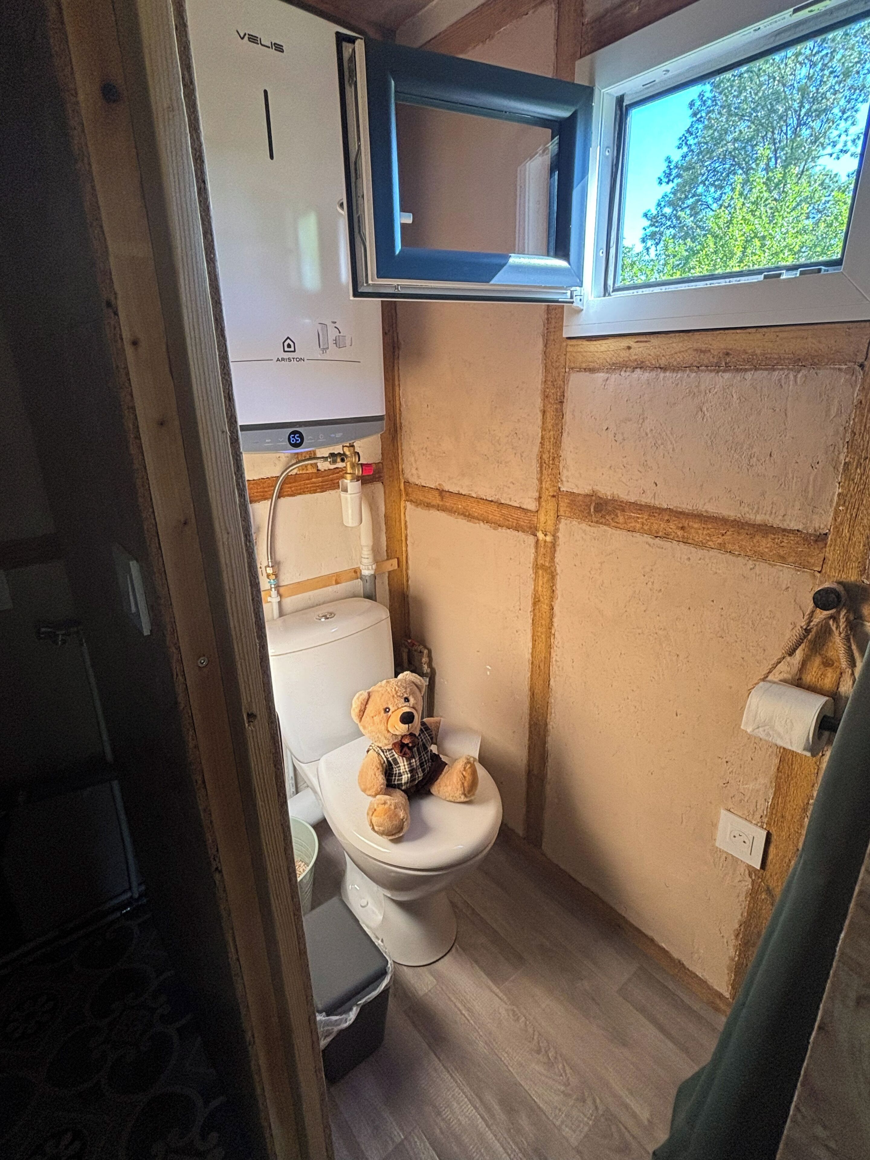 A small bathroom with a toilet, a window, and a bear plush toy on the toilet seat.