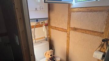 A small bathroom with a toilet, a window, and a bear plush toy on the toilet seat.