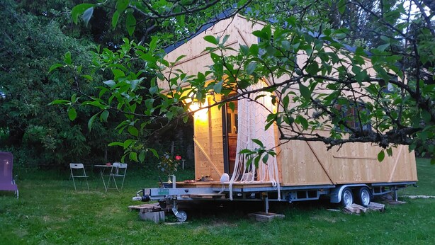A small wooden structure with a flat roof, possibly a cabin or a small house, situated on a grassy area with trees around. - Hempcrete Tiny House (Malbrans)