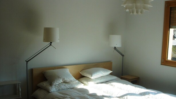 A bedroom with a wooden headboard, two bedside lamps, a window with blinds, and a radiator. - Tgèsa Muntaniala Stamm, (Sedrun), 198.02 by Interhome (Sedrun)